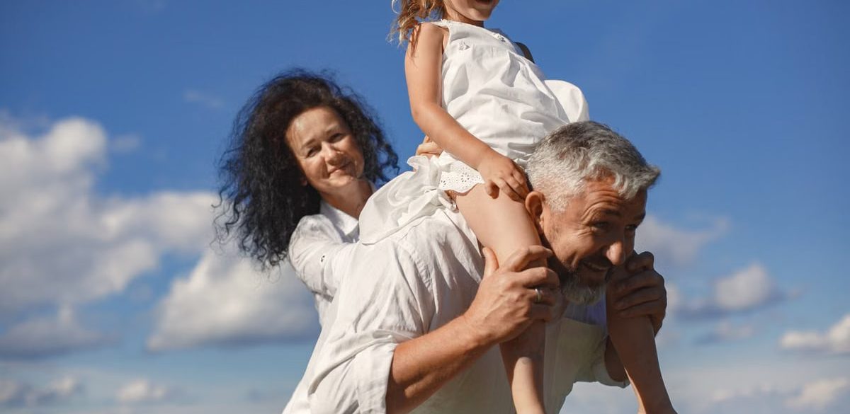 Grandparents playing with granddaughter outdoors under bright blue sky.