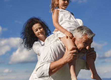 Grandparents playing with granddaughter outdoors under bright blue sky.