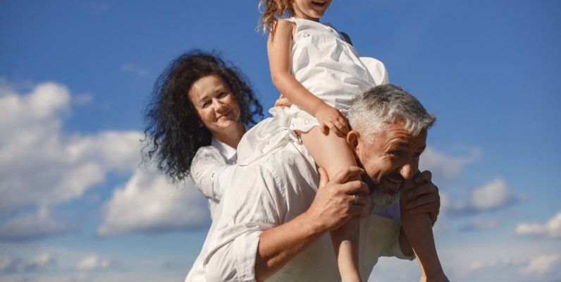 Grandparents playing with granddaughter outdoors under bright blue sky.