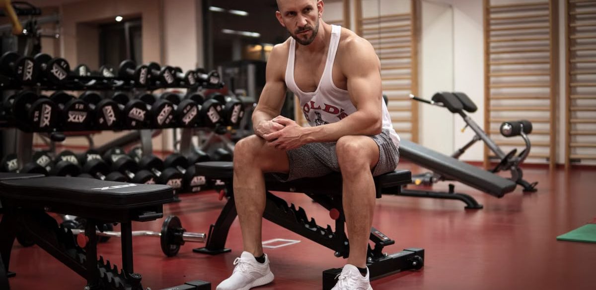 Strong, determined man in white tank top sitting in gym, showcasing fitness and strength.